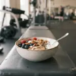 A modest, fresh bowl of natural ingredients like berries, oats, and nuts rests on a clean, dark grey gym bench