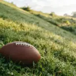 A pristine American football rests at the base of a steep, dew-kissed grassy hill
