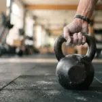 Main de personne tenant un kettlebell en fer posé sur le sol d'une salle de sport